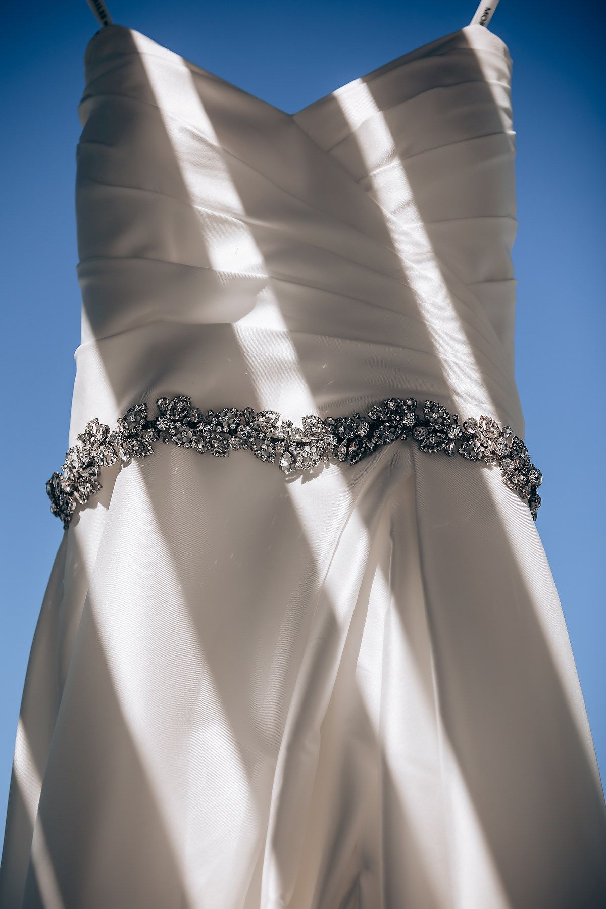 Close-up details of an elegant white wedding dress with beaded embellishments against a blue sky background.