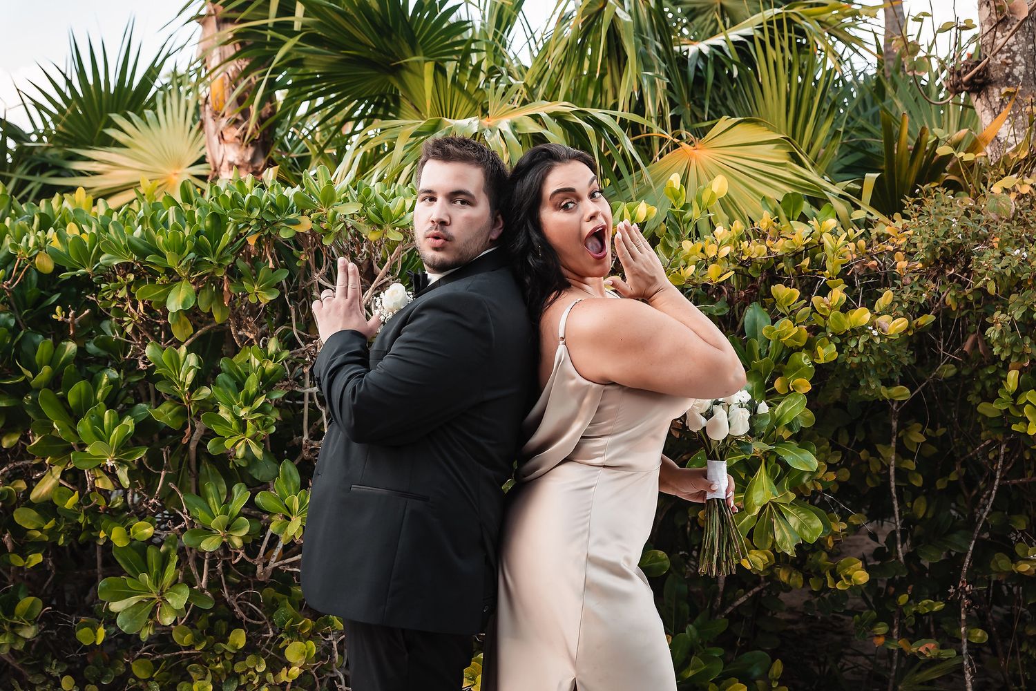 Wedding party poses for photos outdoors with tropical palm trees and greenery in the background.