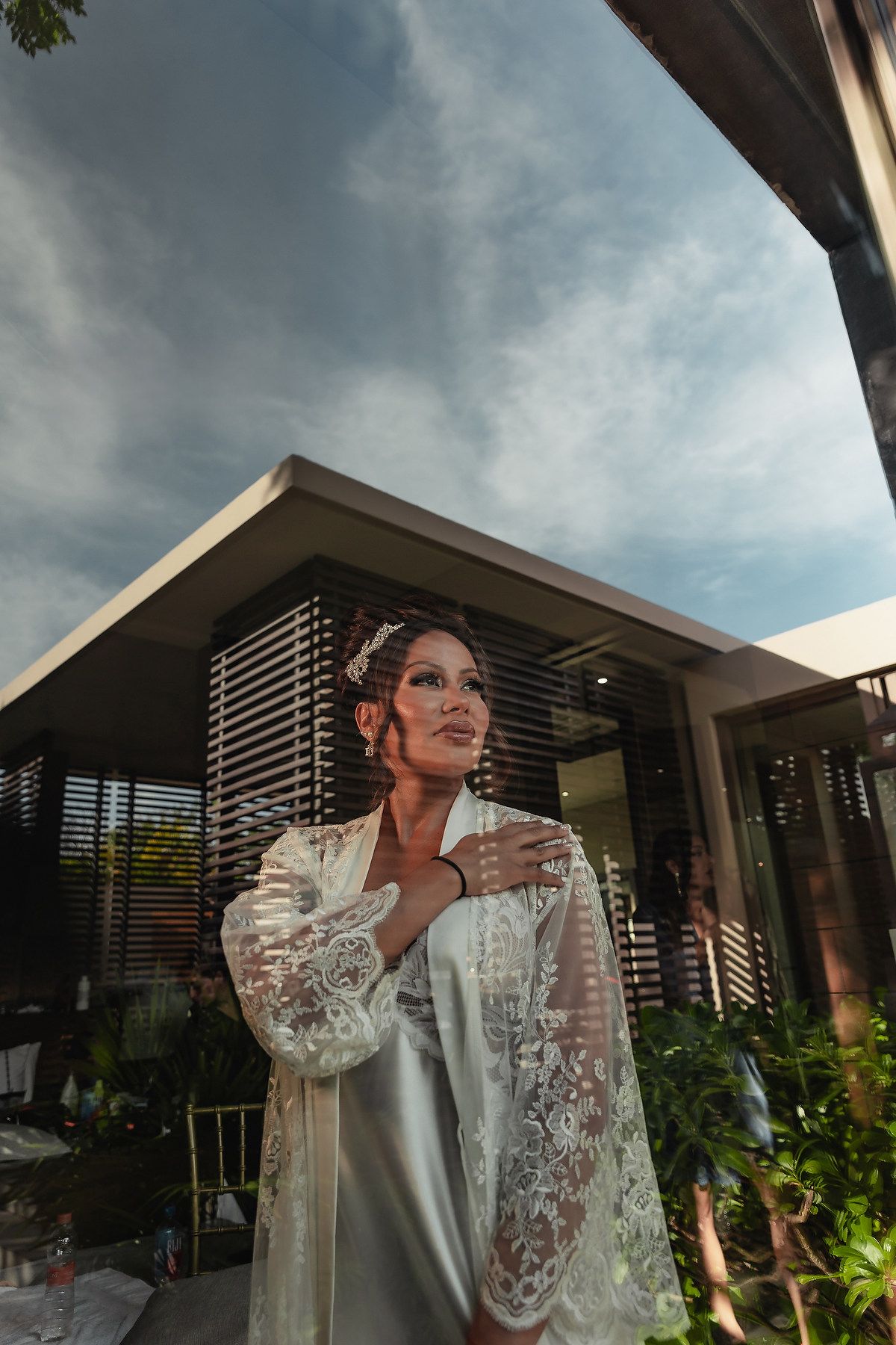 A person in a white lace dress poses dramatically against a modern building exterior with tropical landscaping at sunset.