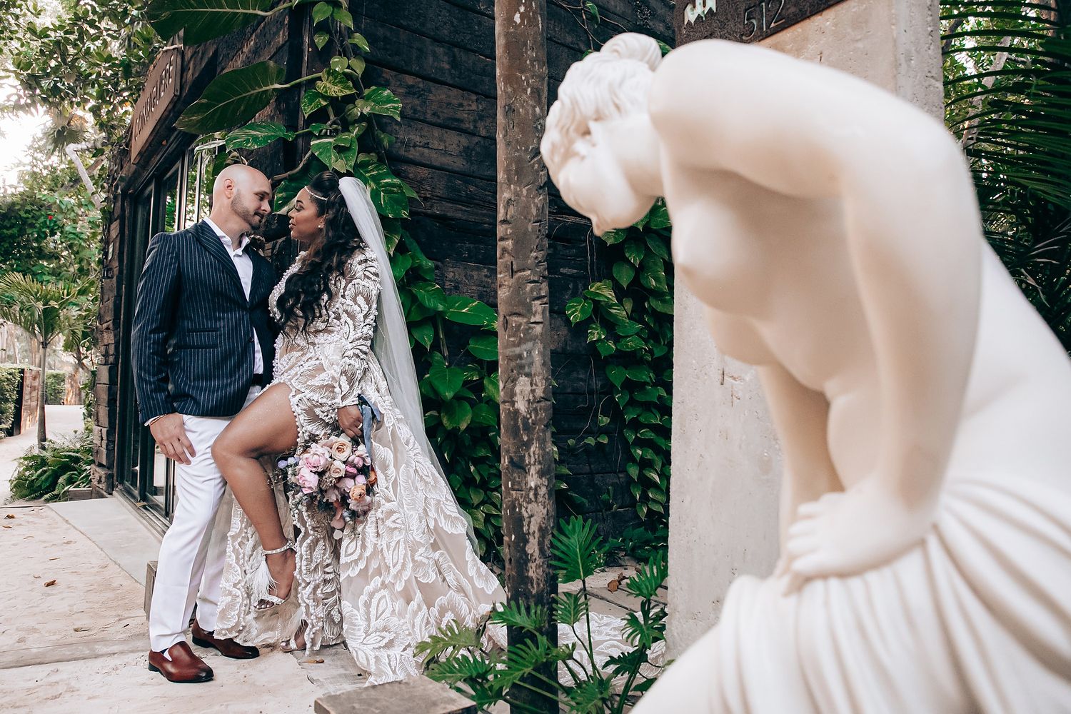 A romantic outdoor wedding scene with a couple embracing under tall palm trees in a garden setting.