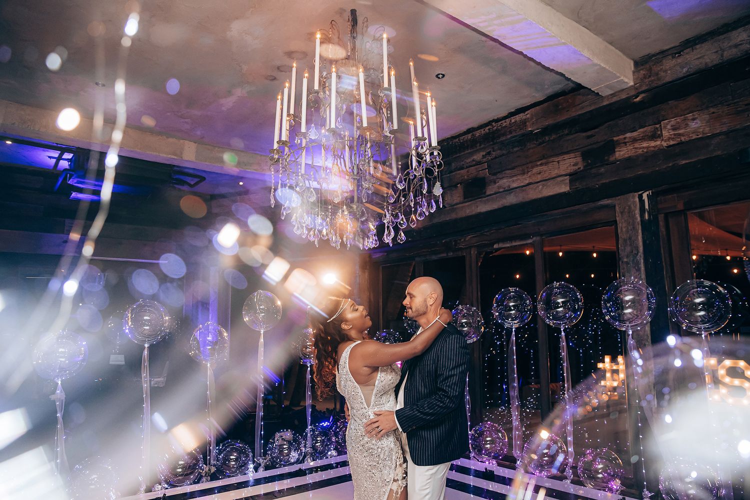 A couple shares a romantic dance moment under elegant chandeliers at an evening wedding reception.