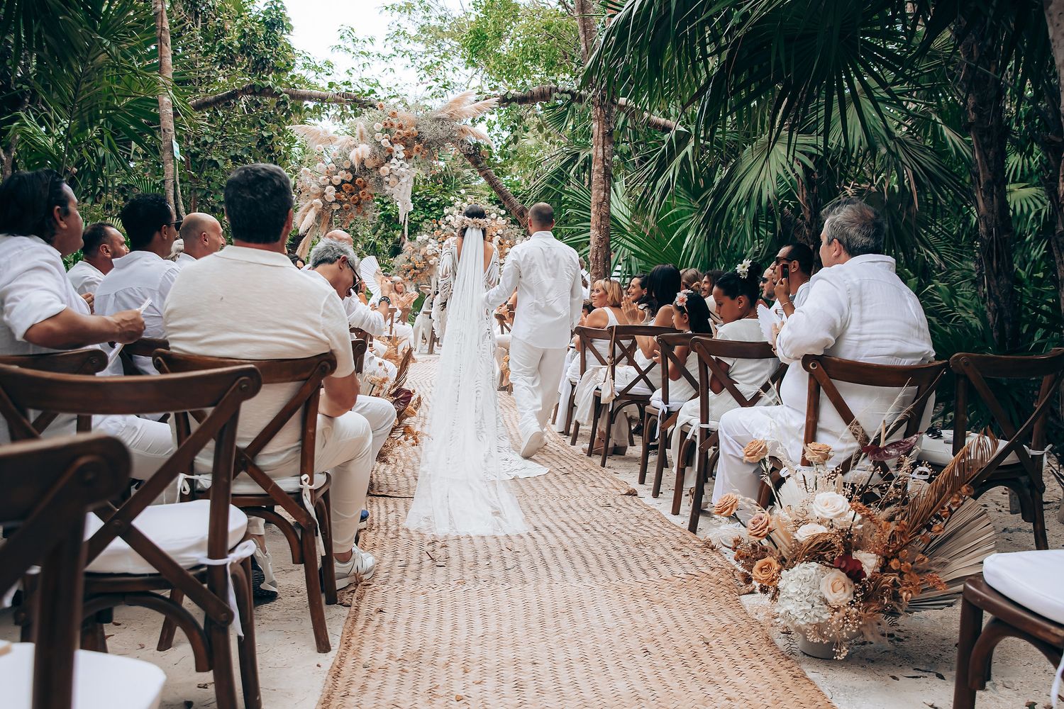 Tropical beach wedding ceremony setup with wooden chairs and palm trees along a sandy aisle.