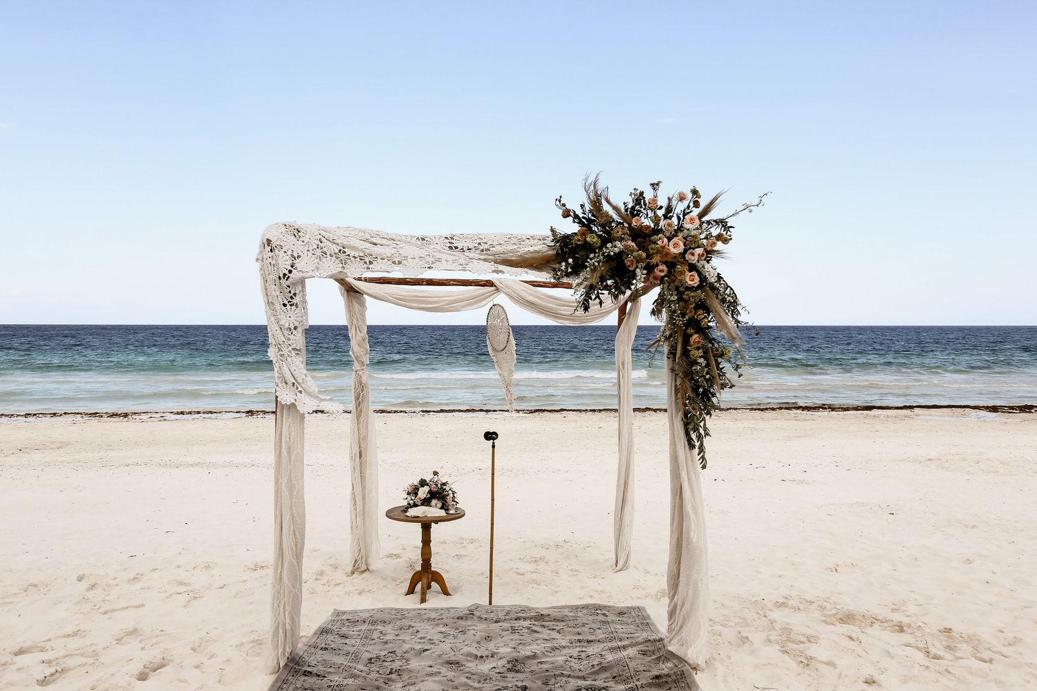 Minimalist beach wedding arch with white fabric and dried flower arrangements on sandy shore.
