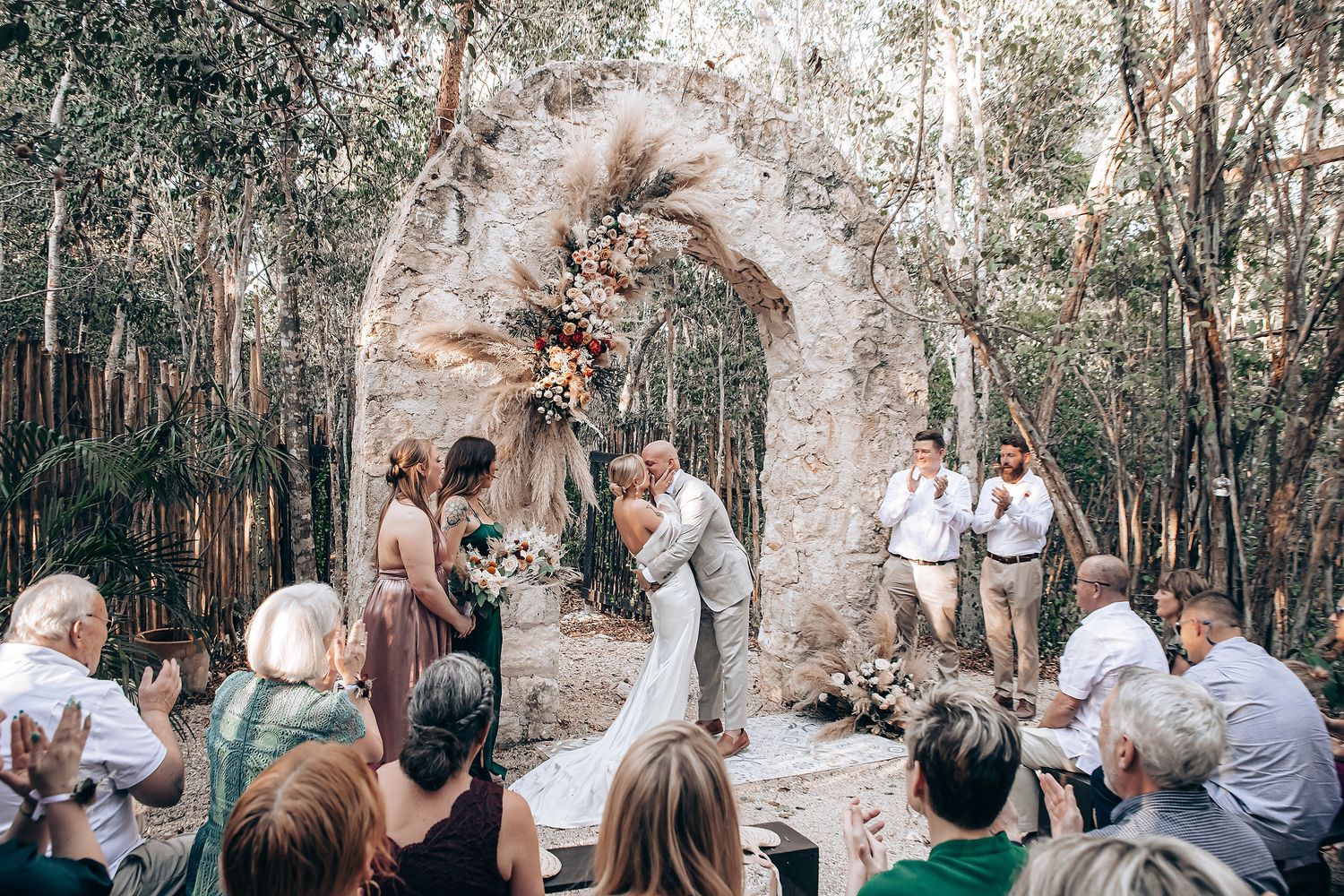 Intimate outdoor wedding ceremony under a rustic stone arch surrounded by trees and guests.