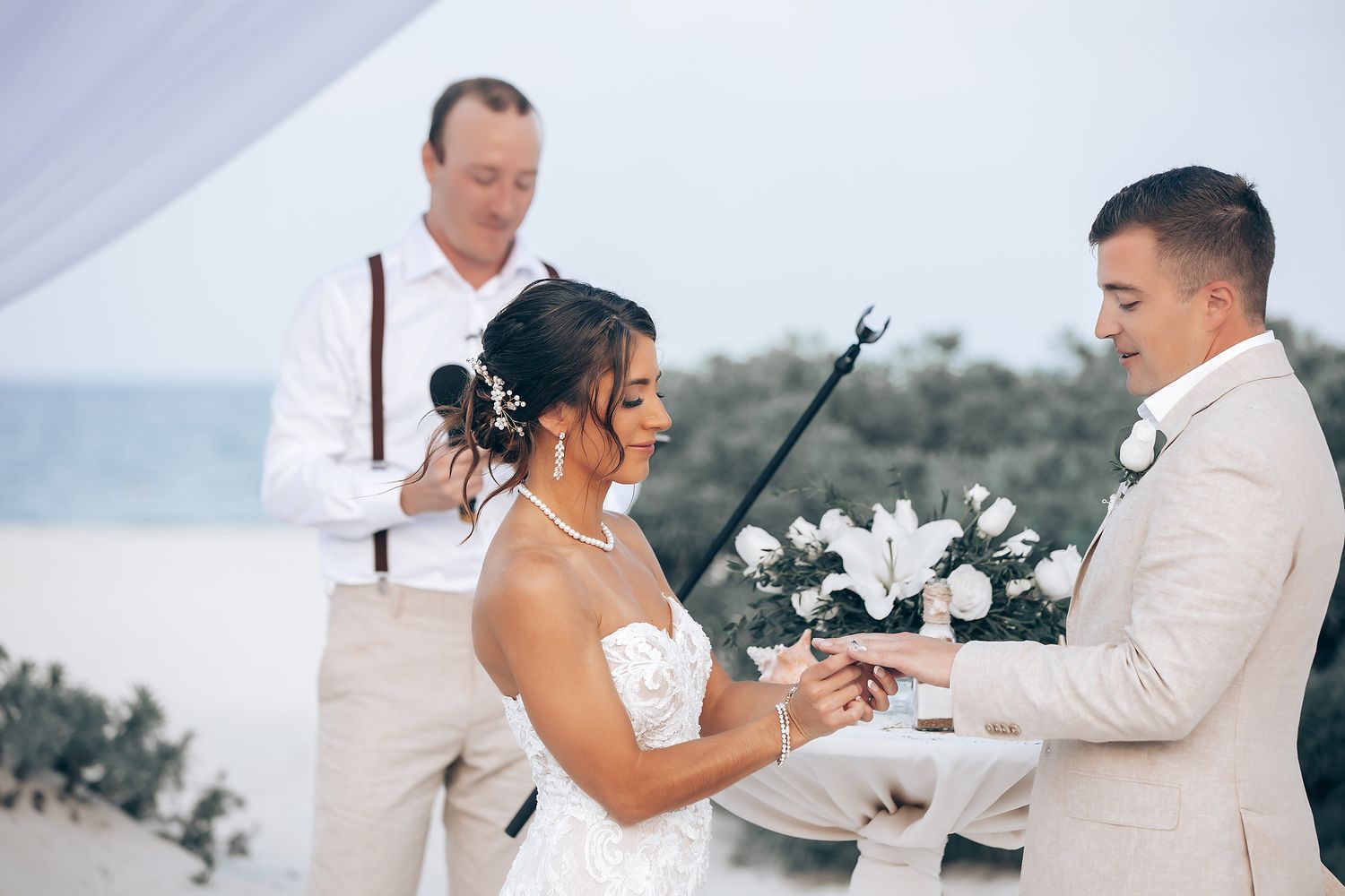 Exchange of rings during beachfront wedding ceremony under white draping.