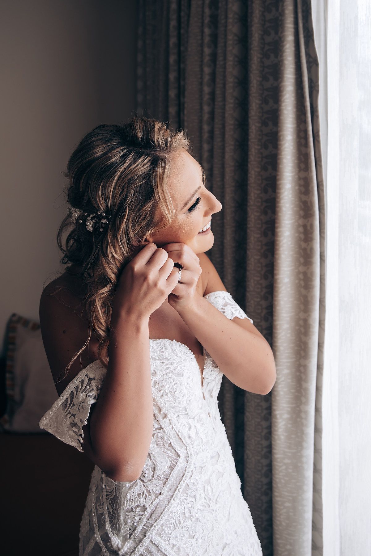 Bride in a white lace wedding dress puts on earrings while getting ready by a window with grey curtains.
