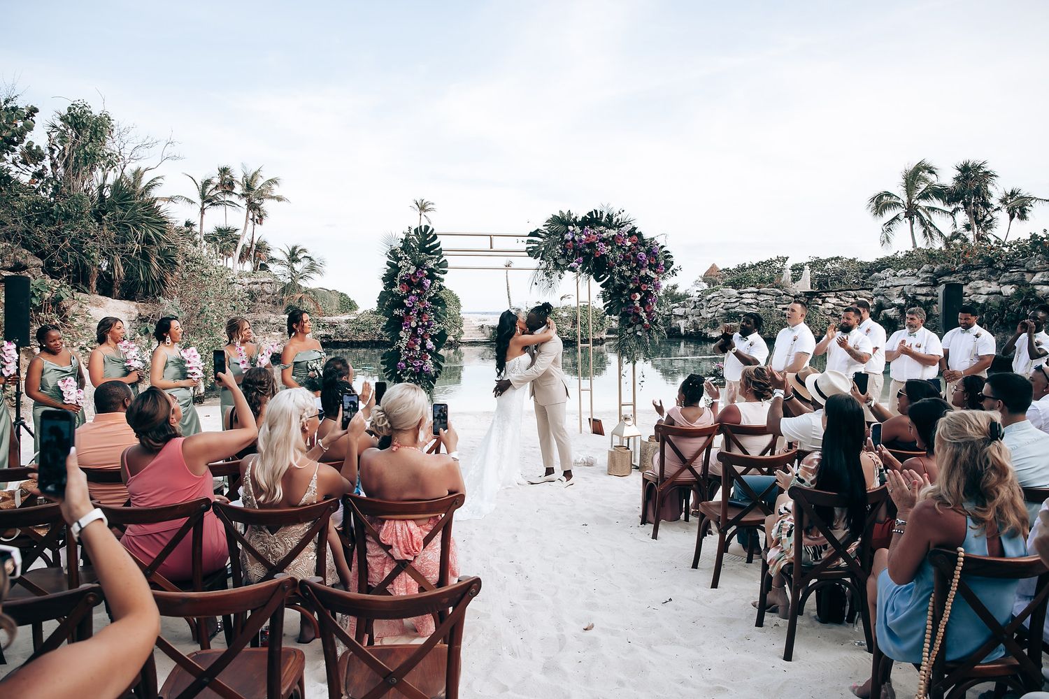 Wedding guests seated in wooden chairs watching beachside ceremony.