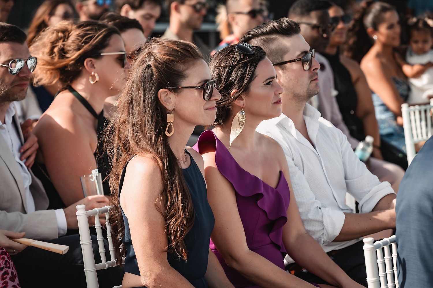 Wedding guests in summer attire watching ceremony from white chairs.