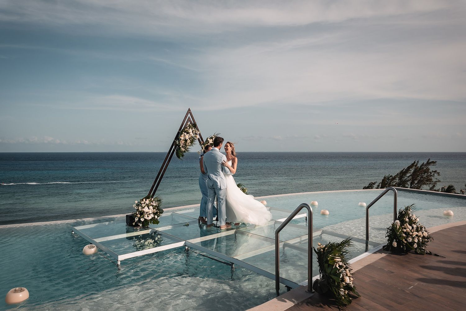 Romantic wedding ceremony on oceanfront deck with floral triangle arch and infinity pool.