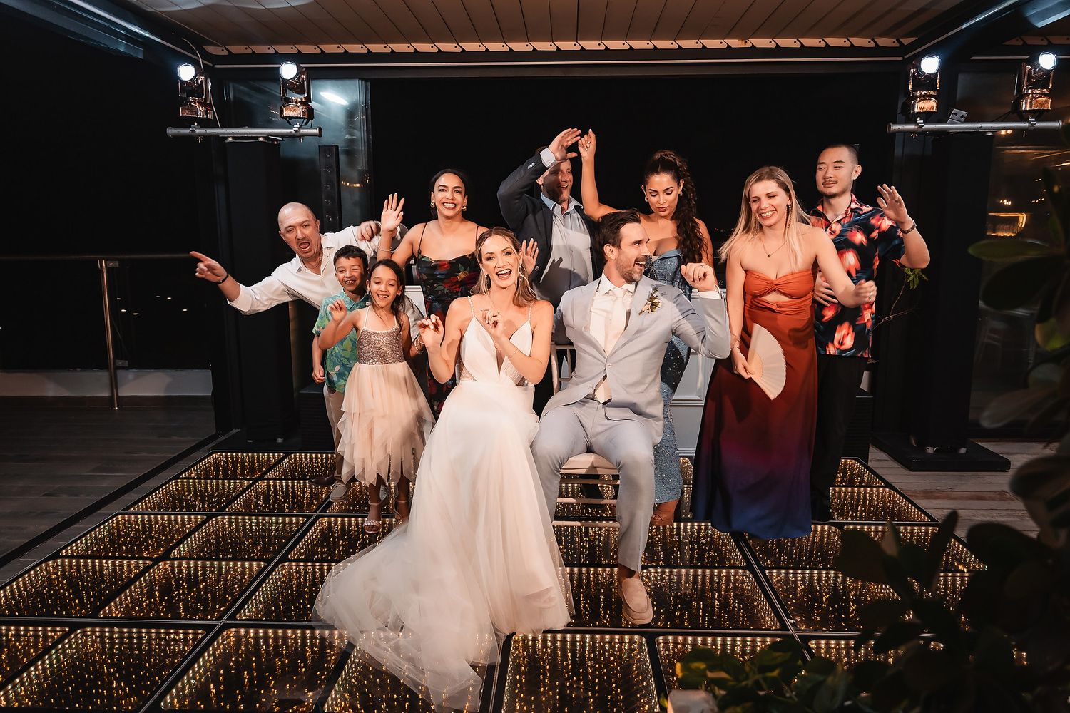 Wedding celebration group photo with party striking fun poses on lit-up dance floor.