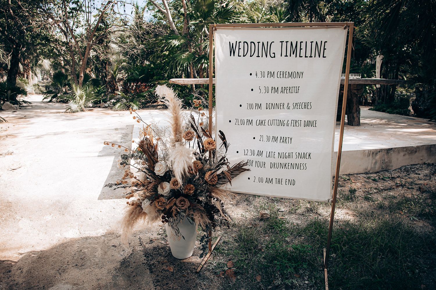 A rustic wedding timeline sign displayed outdoors with dried floral arrangements and natural lighting.