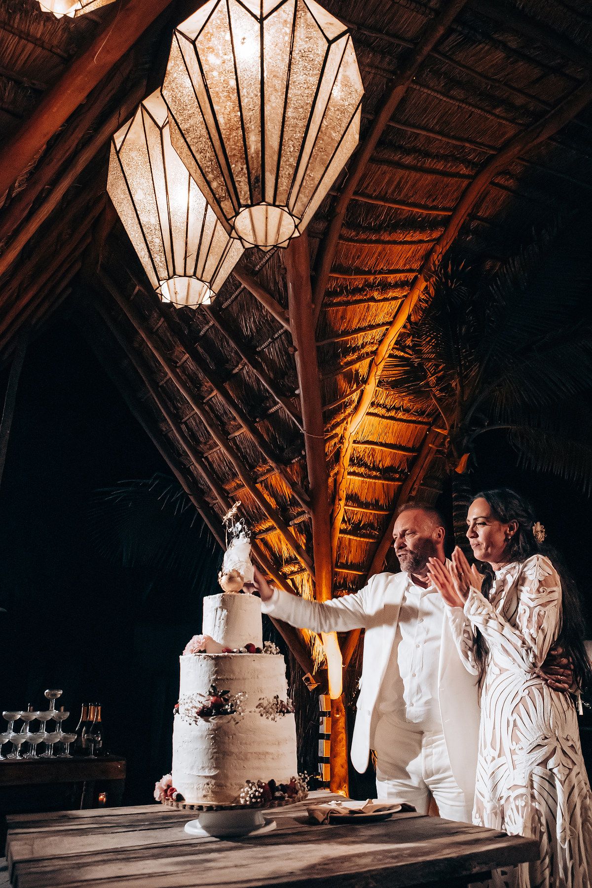 A wedding couple in white attire admires their elegant three-tiered cake in a rustic wooden venue with pendant lighting.