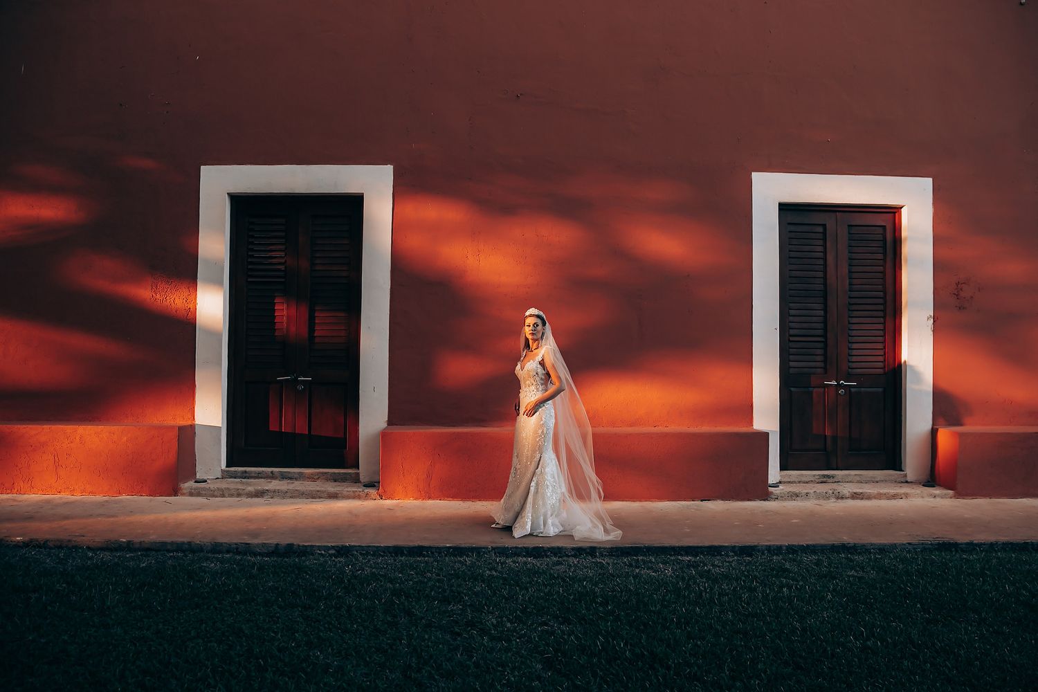 A bride in a flowing white dress stands against a dramatic red sunset sky and white building facade.