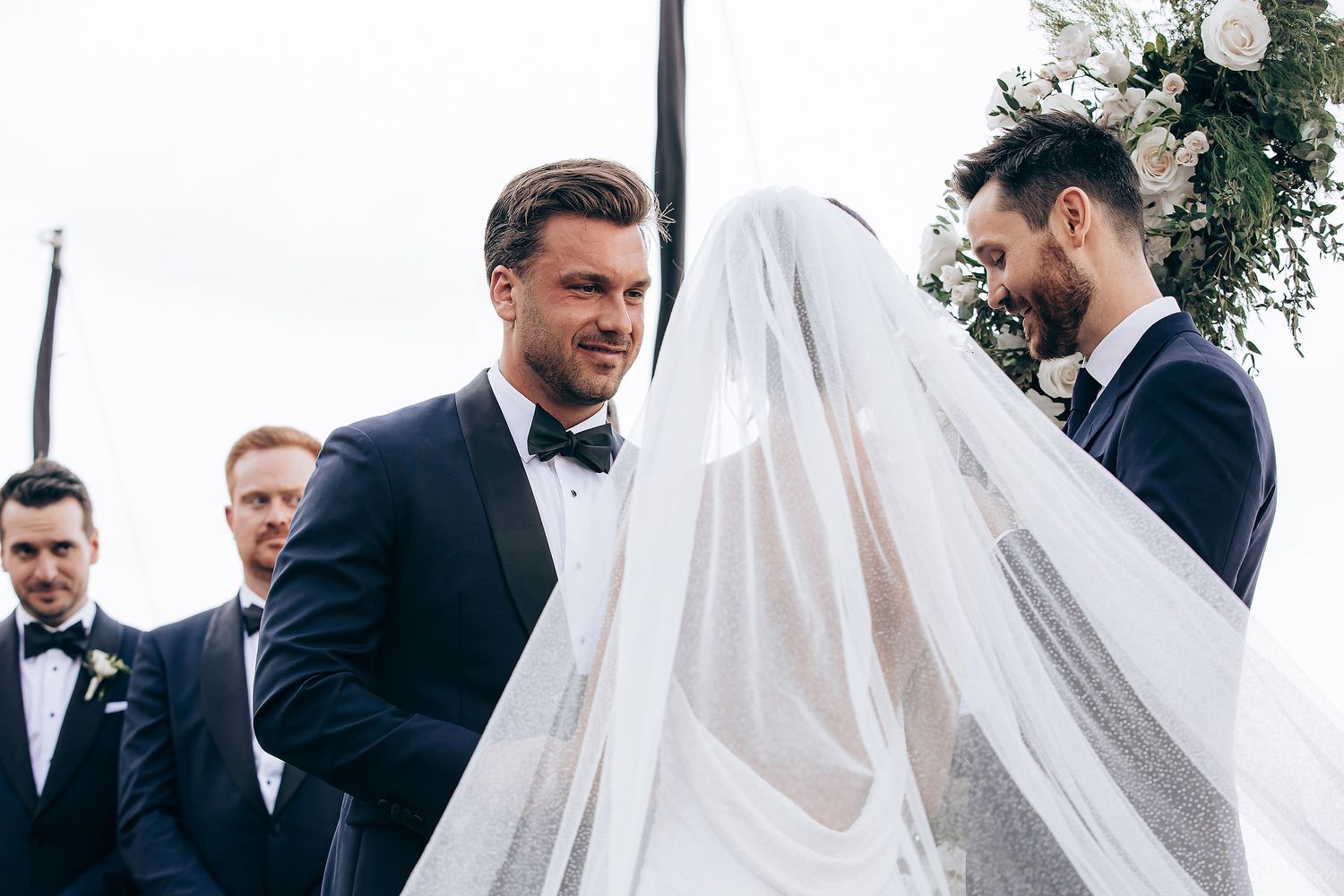 A wedding party in formal attire gathers outdoors with flowing white veil creating an ethereal moment.