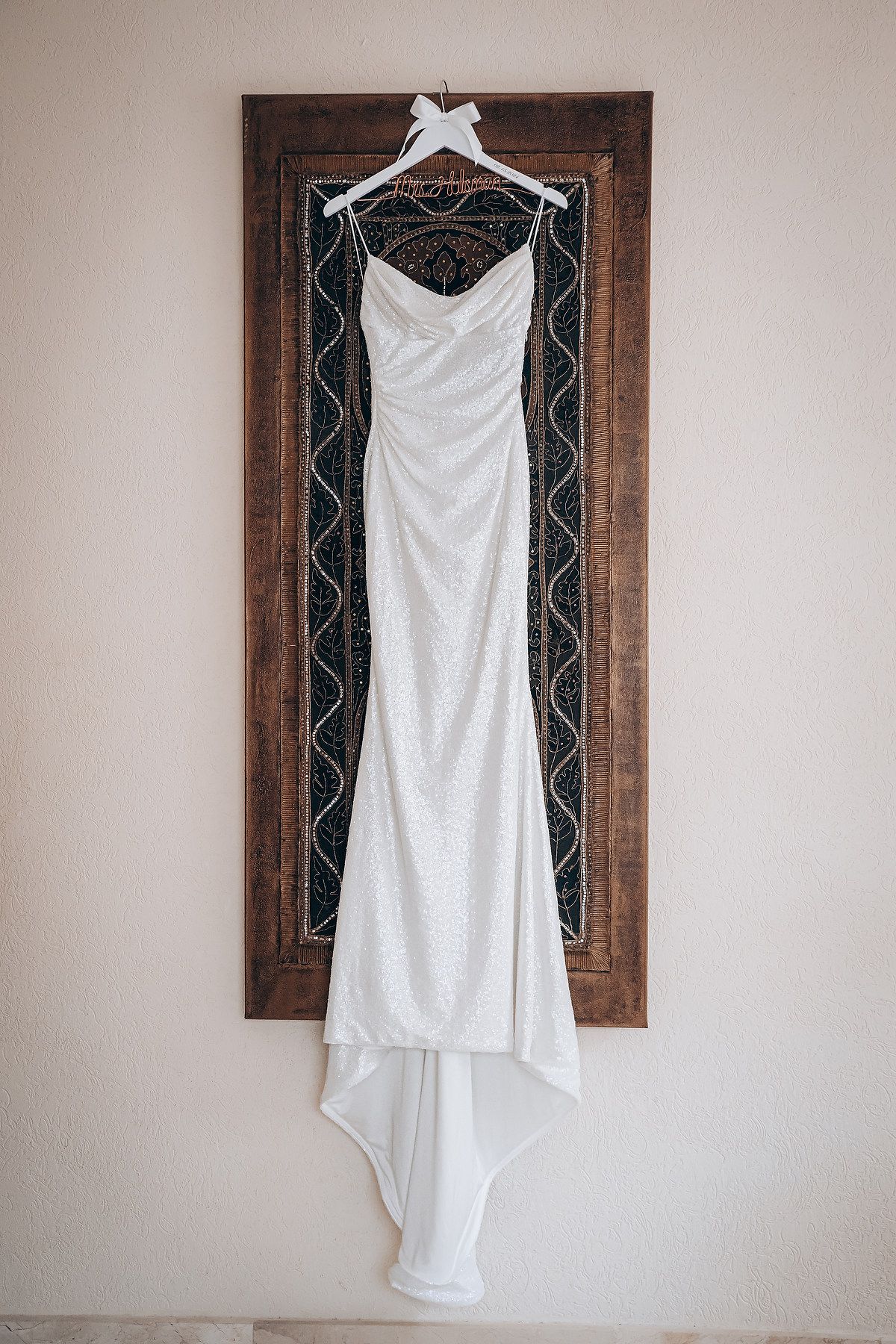 Elegant white wedding dress displayed on a hanger against a wall with decorative wooden framed artwork.