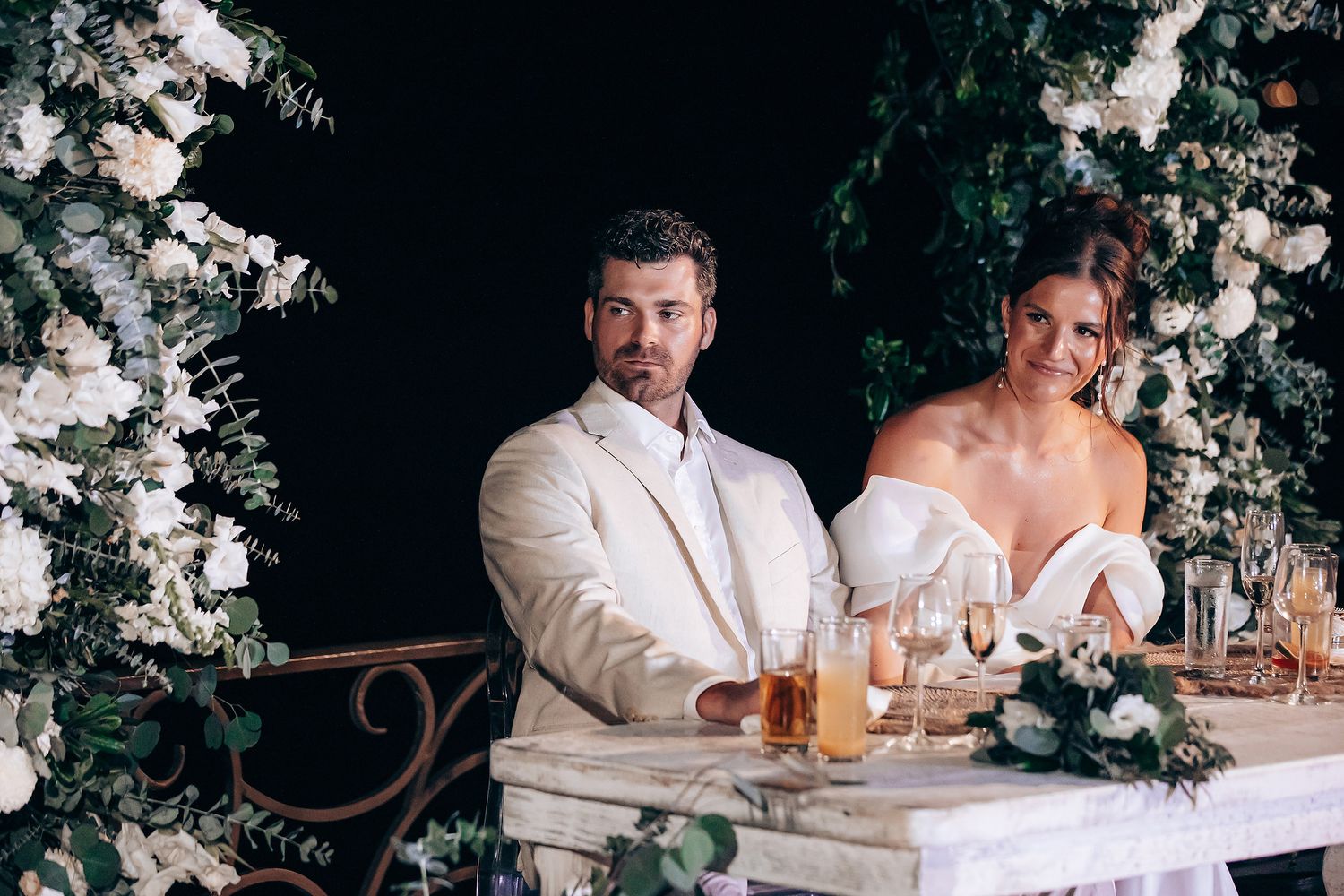 A couple sits at an elegant wedding reception table decorated with white floral arrangements and candlelight.
