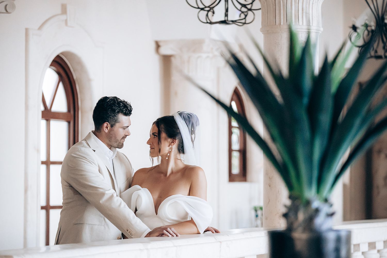 A romantic couple shares a tender moment in an elegant white interior with arched windows and a decorative plant.