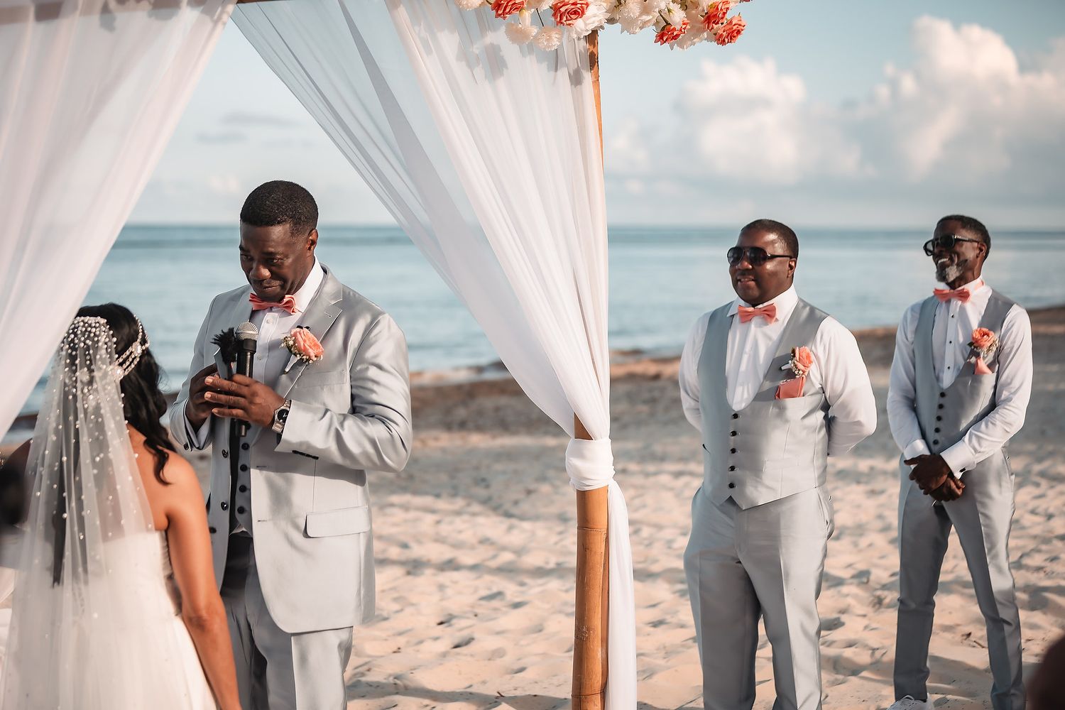 Beachfront wedding ceremony with white drapes, coral flowers, and guests in formal attire.