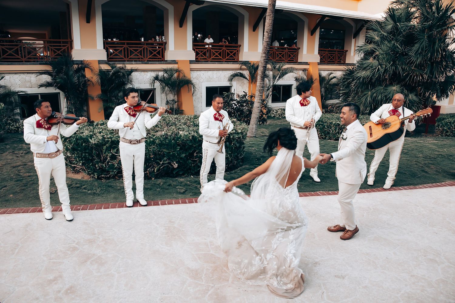 Wedding party members in white attire pose playfully in a line along a Mediterranean-style courtyard.