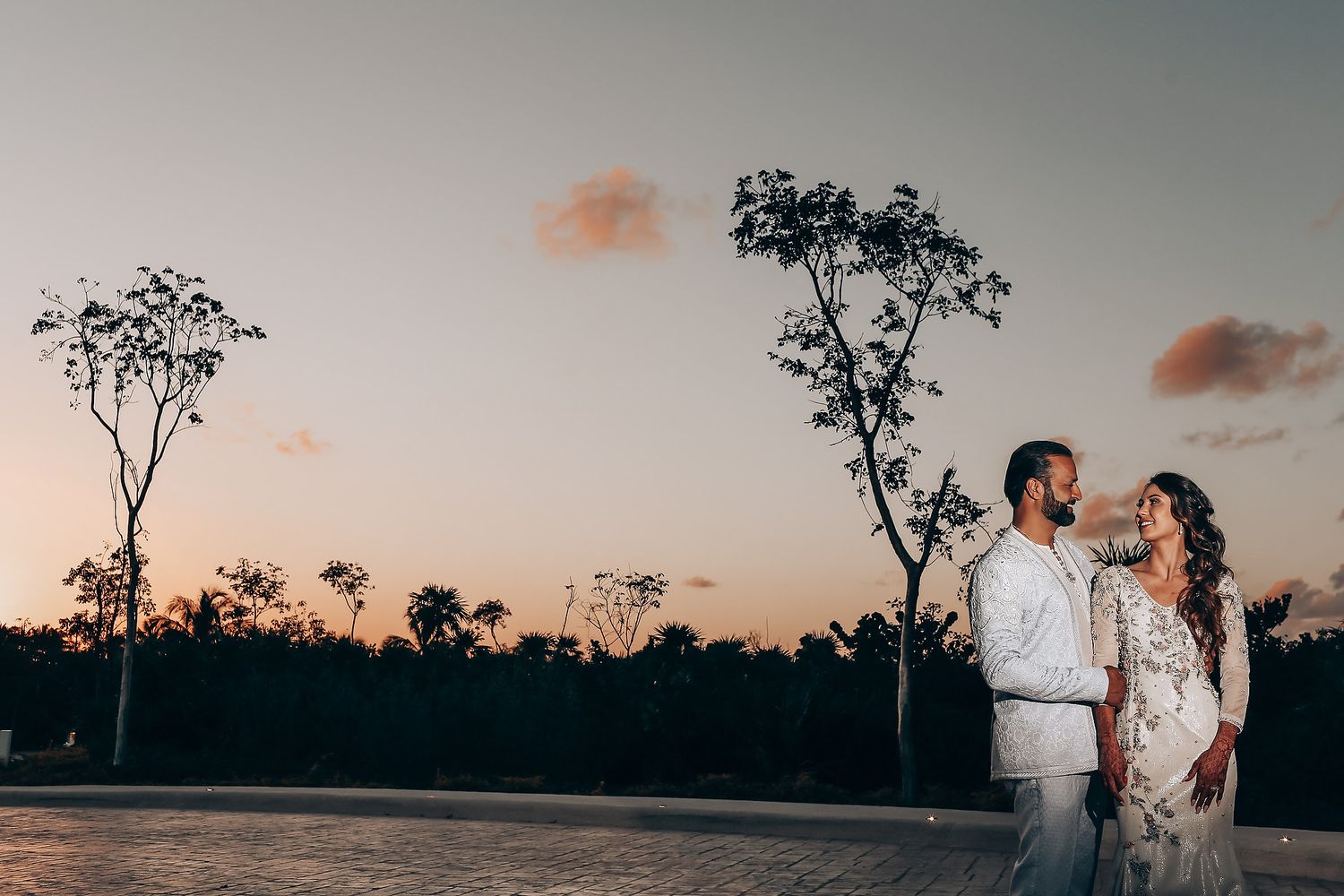 A romantic couple stands silhouetted against a sunset sky with trees in the background at an outdoor location.