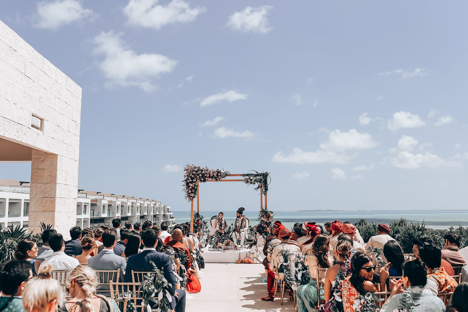 Guests gather at an outdoor beach wedding ceremony with white chairs and ocean views on a sunny day.