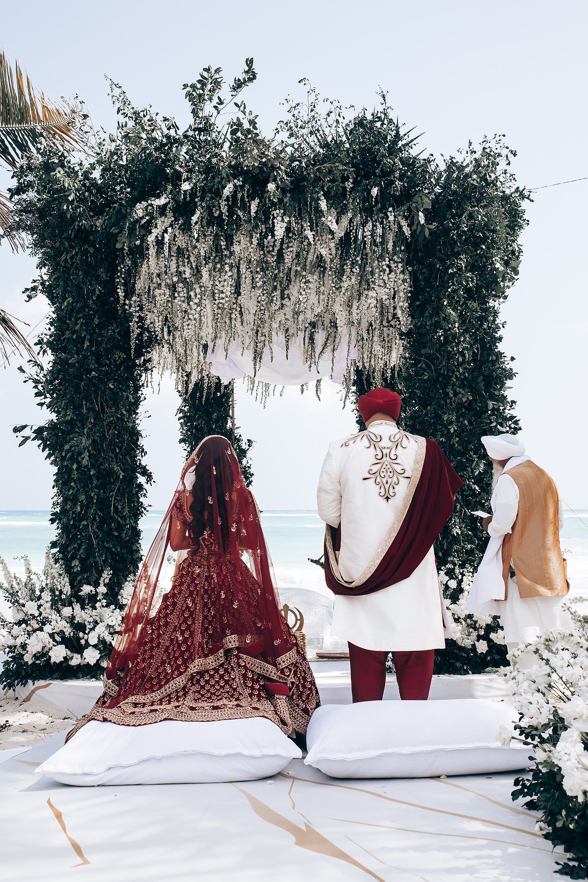 Wedding couple sits on white fabric under floral arch with ocean view.