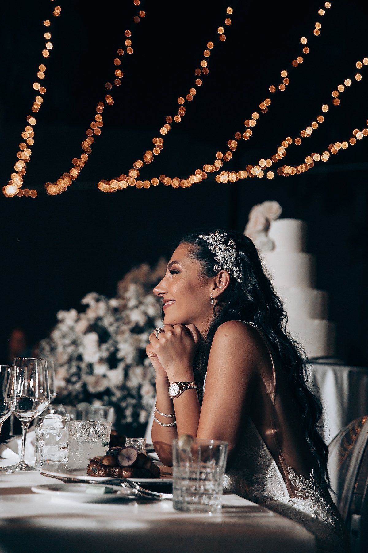 Reception table setting with string lights overhead and wedding cake in background.