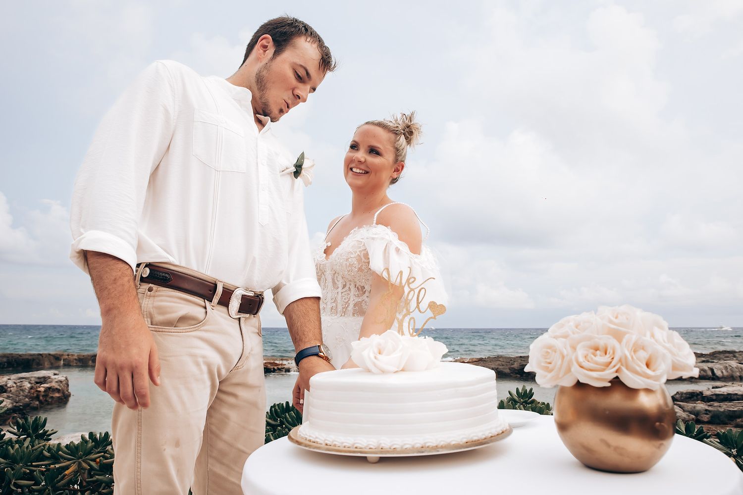 Wedding cake cutting ceremony with ocean view and tropical backdrop.