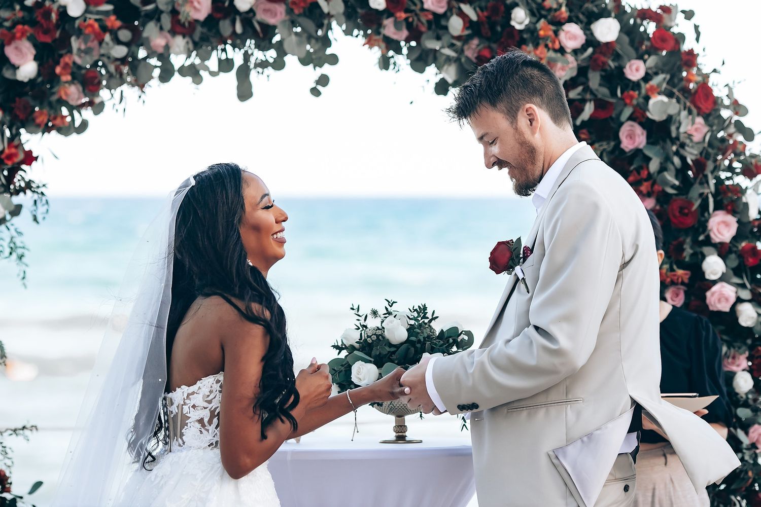 A romantic beach wedding ceremony under a floral arch with pink and red roses by the ocean.