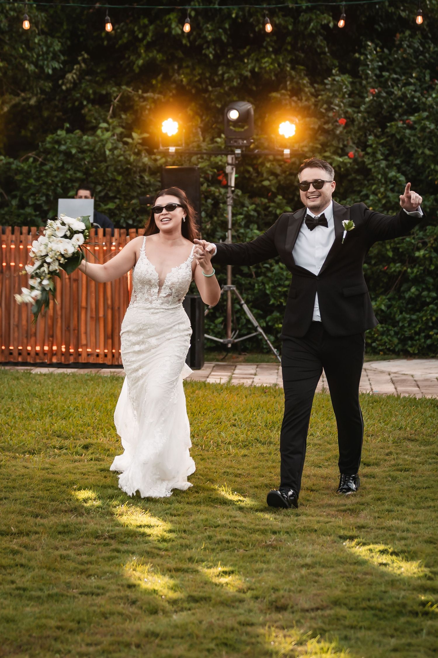 Wedding couple dances playfully on grass under string lights at outdoor reception.
