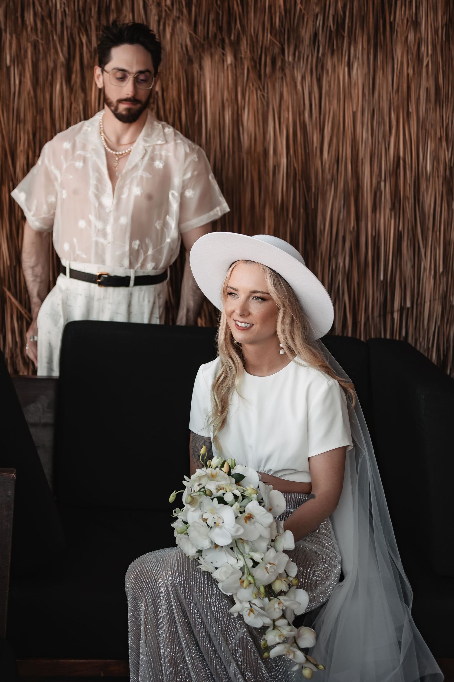 Wedding couple posing against a rustic thatched wall backdrop with white floral bouquet.