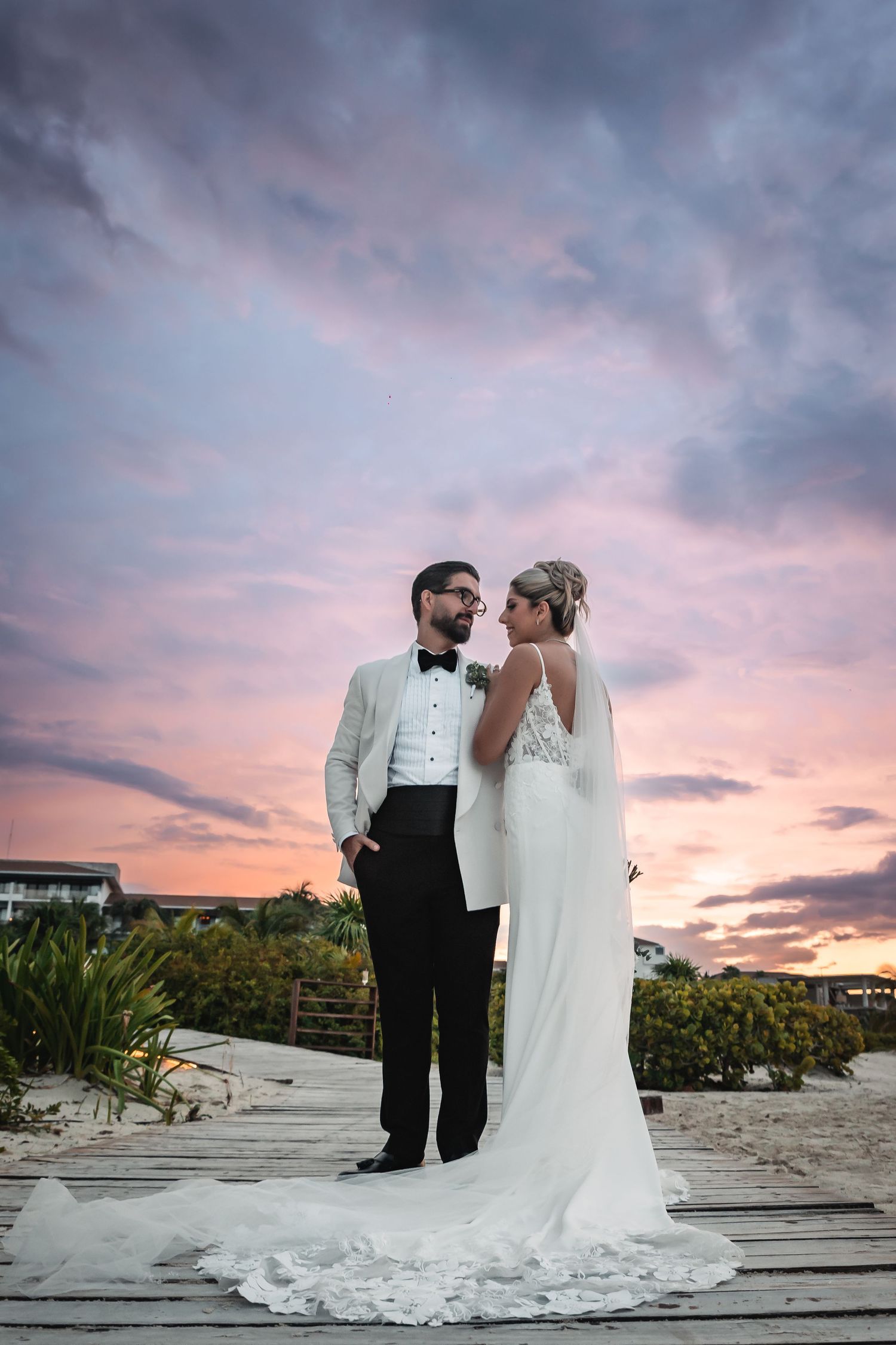 Wedding couple poses on wooden boardwalk against dramatic pink sunset sky.