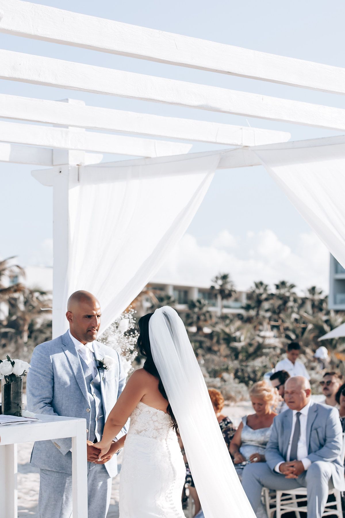 Elegant beach wedding ceremony under a white pergola with guests seated on a sunny day.