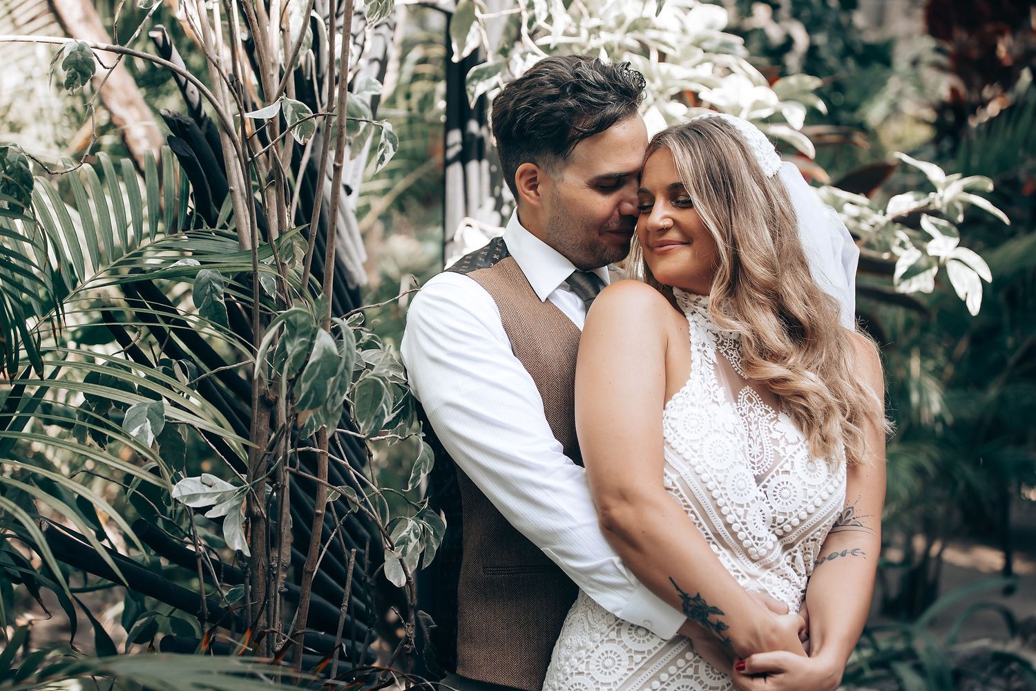 A romantic couple embraces in a lush tropical garden setting, the bride wearing a white lace dress.