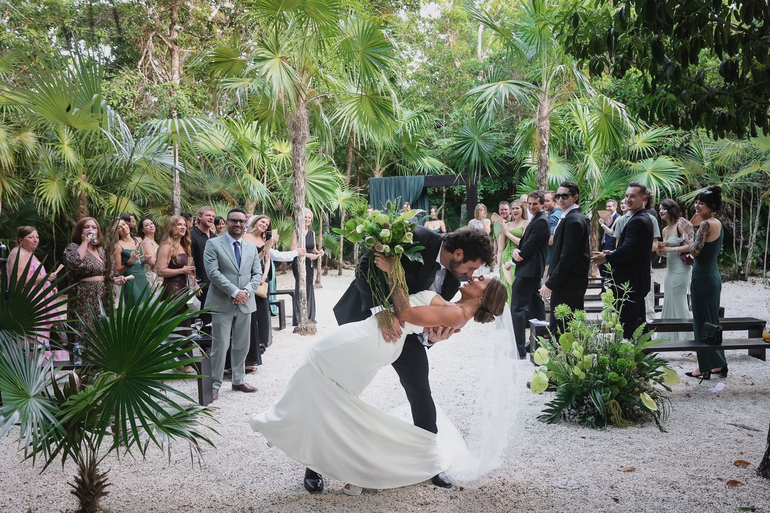 Groom dips bride at outdoor tropical wedding ceremony surrounded by lush palm trees and guests.