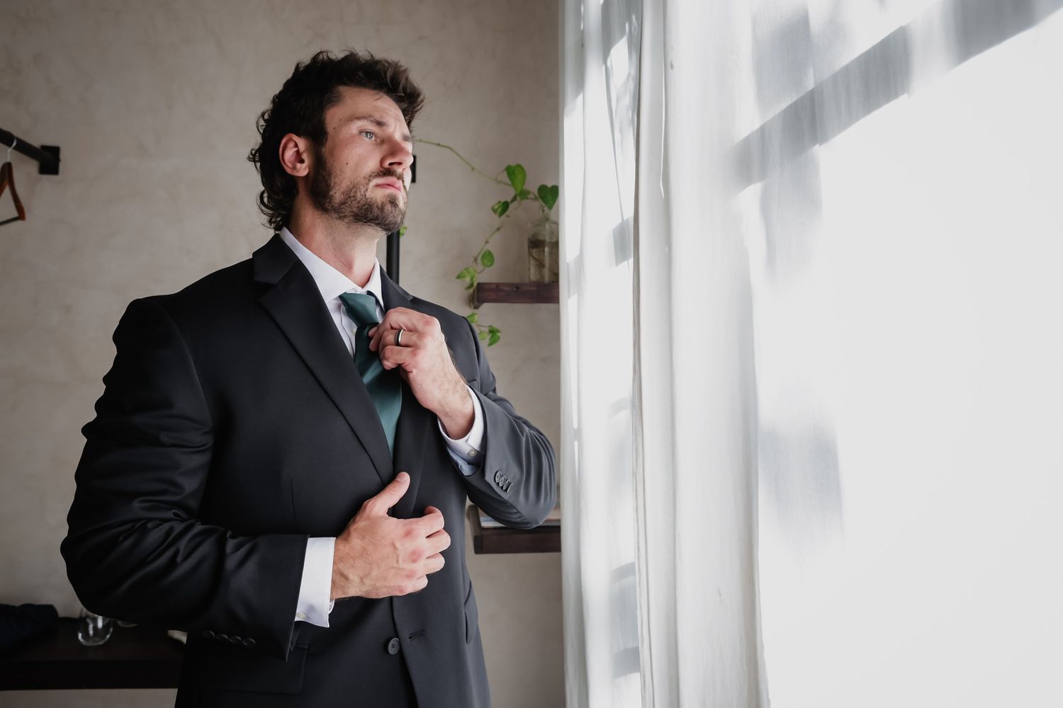 Elegant groom in a dark suit adjusting his tie near a window while preparing for his wedding ceremony.
