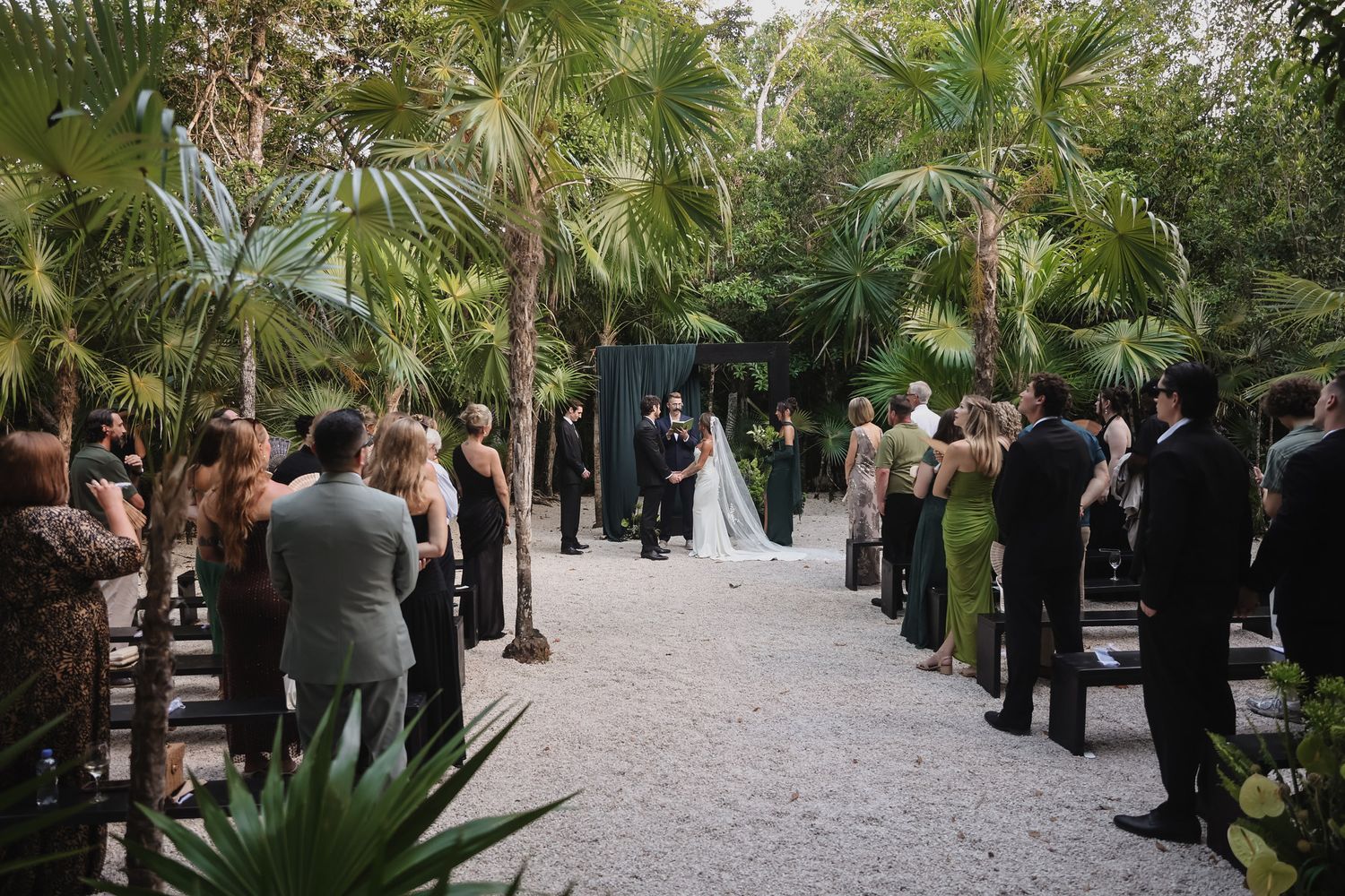 Outdoor wedding ceremony in lush tropical garden with guests gathered along sandy aisle.