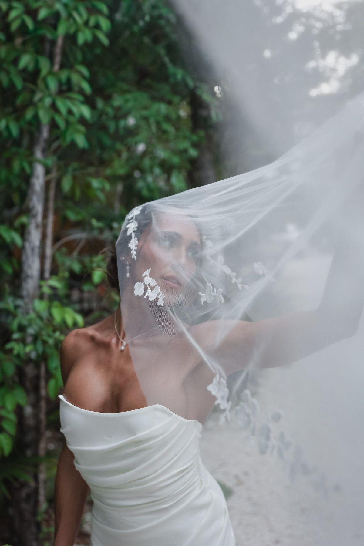 Bride in white strapless gown with floral veil blowing over her face, posing outdoors among lush green trees.