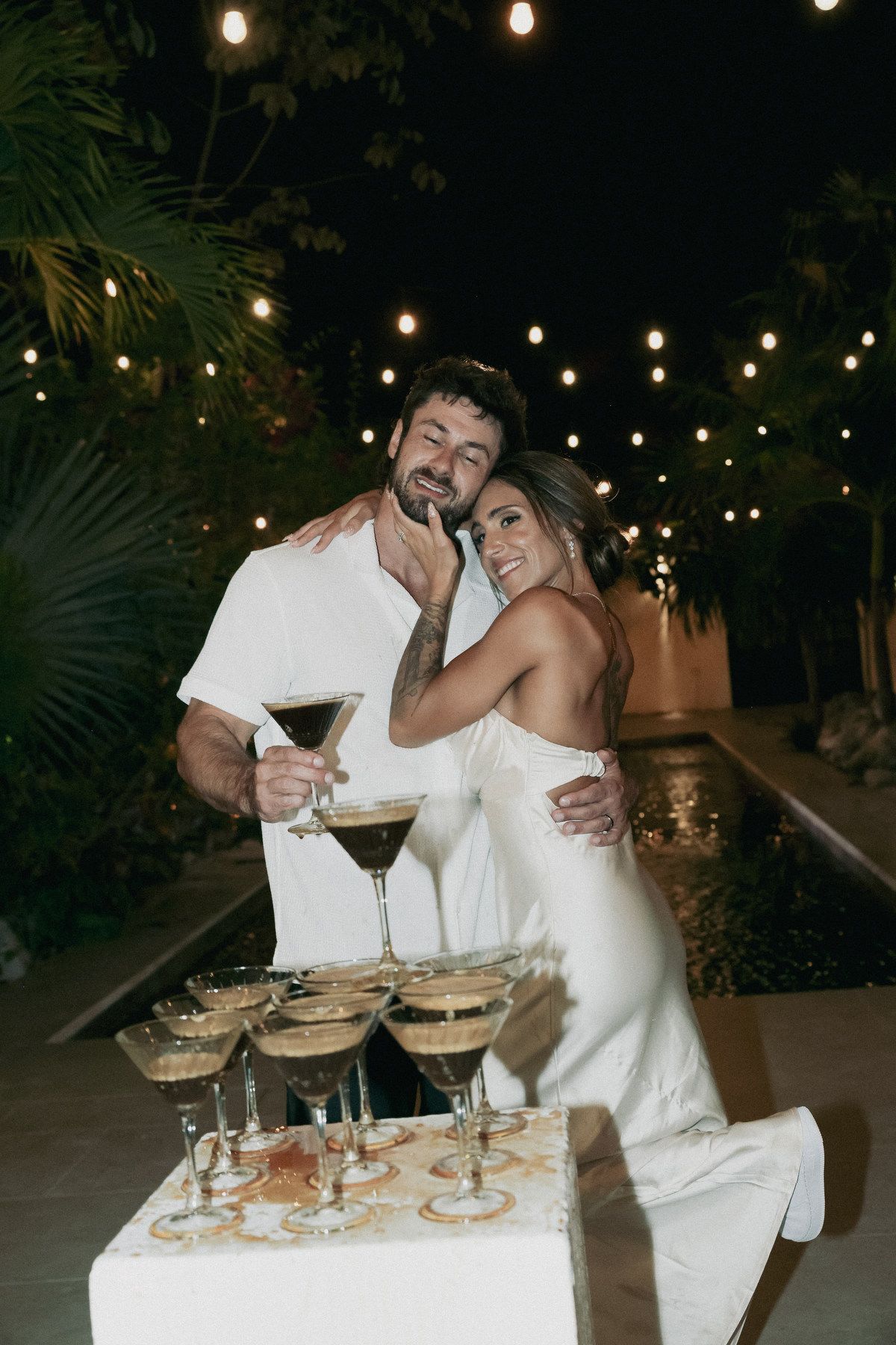 Couple posing with espresso martini tower at outdoor evening wedding reception with string lights.