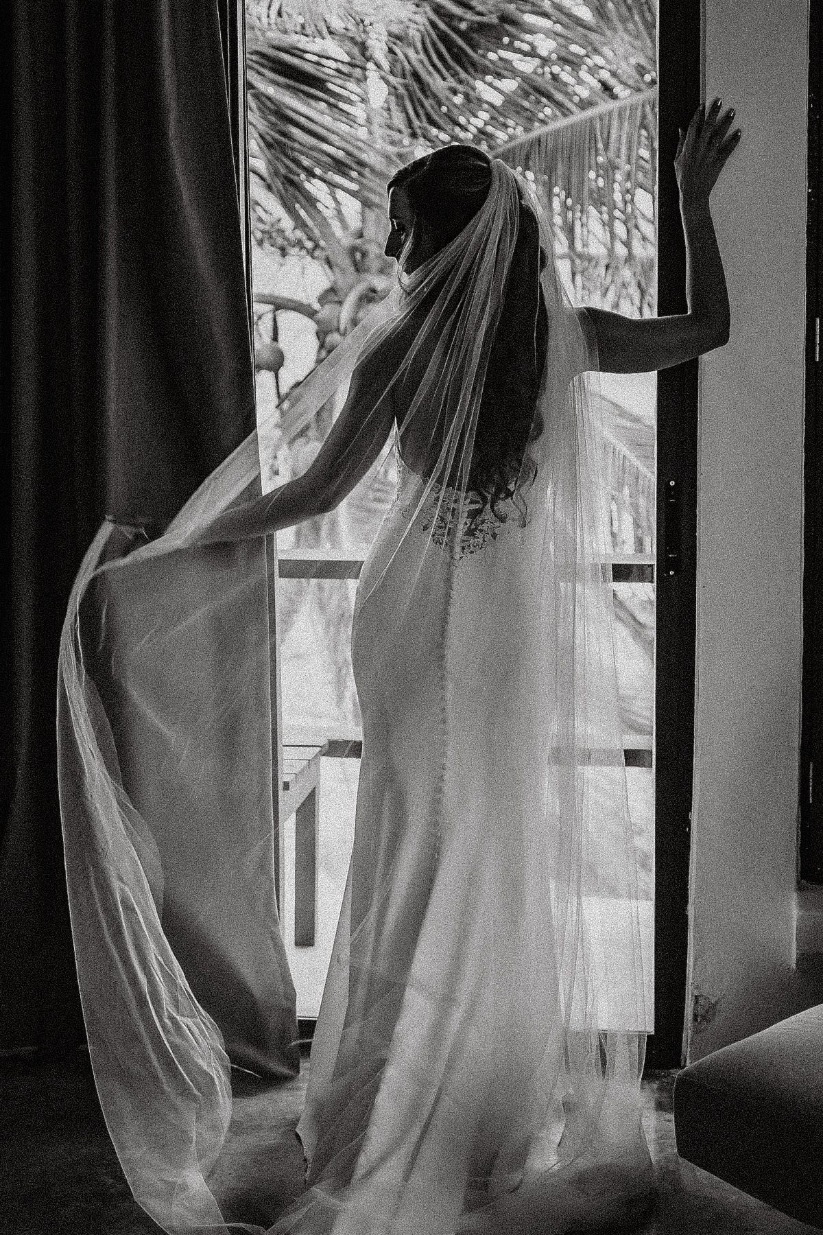 Black and white photo of a bride in a wedding dress and long veil standing by an open door with tropical palms.