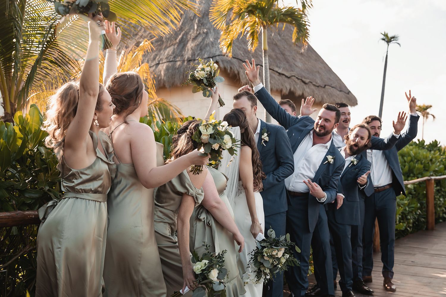 Wedding party celebrating outdoors at tropical venue, bridesmaids in sage green gowns and groomsmen in navy suits cheering joyfully.