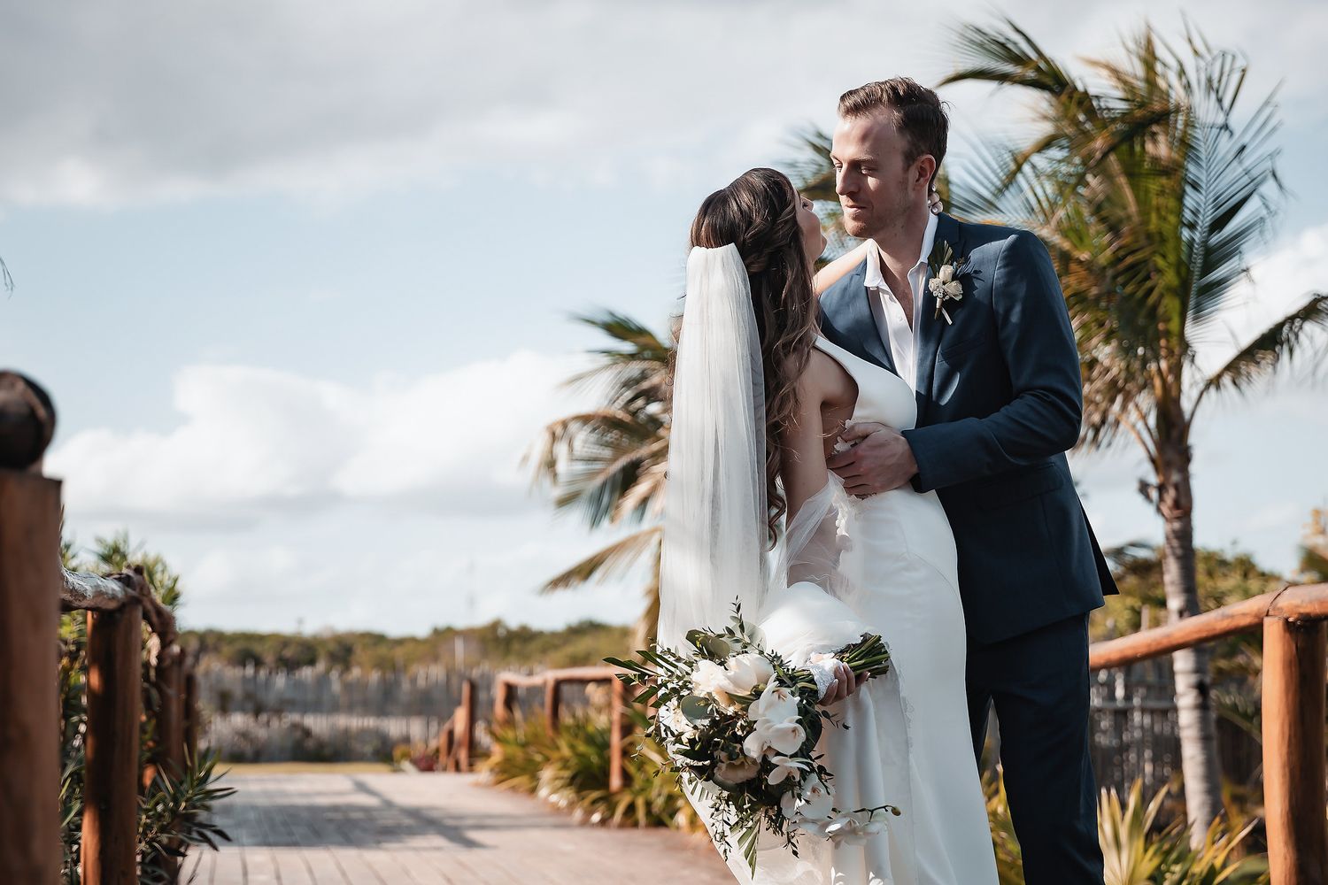Bride and groom embrace on tropical boardwalk, palm trees swaying, bride holding white floral bouquet in wedding attire.