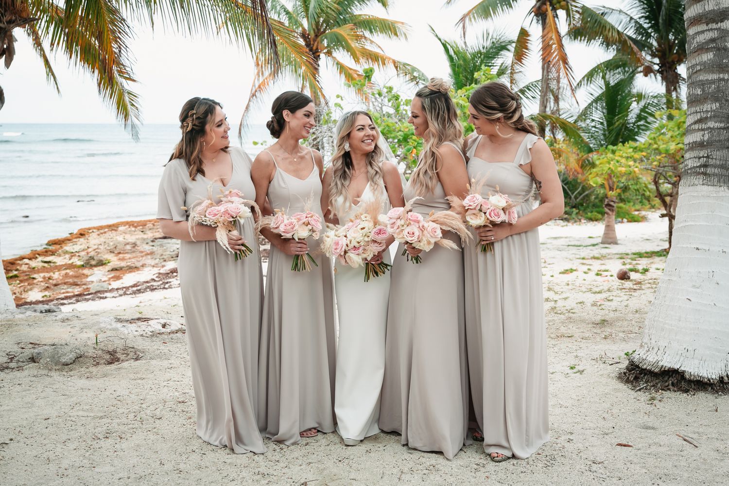 Four bridesmaids in taupe chiffon gowns hold pink bouquets on a tropical beach with palm trees.