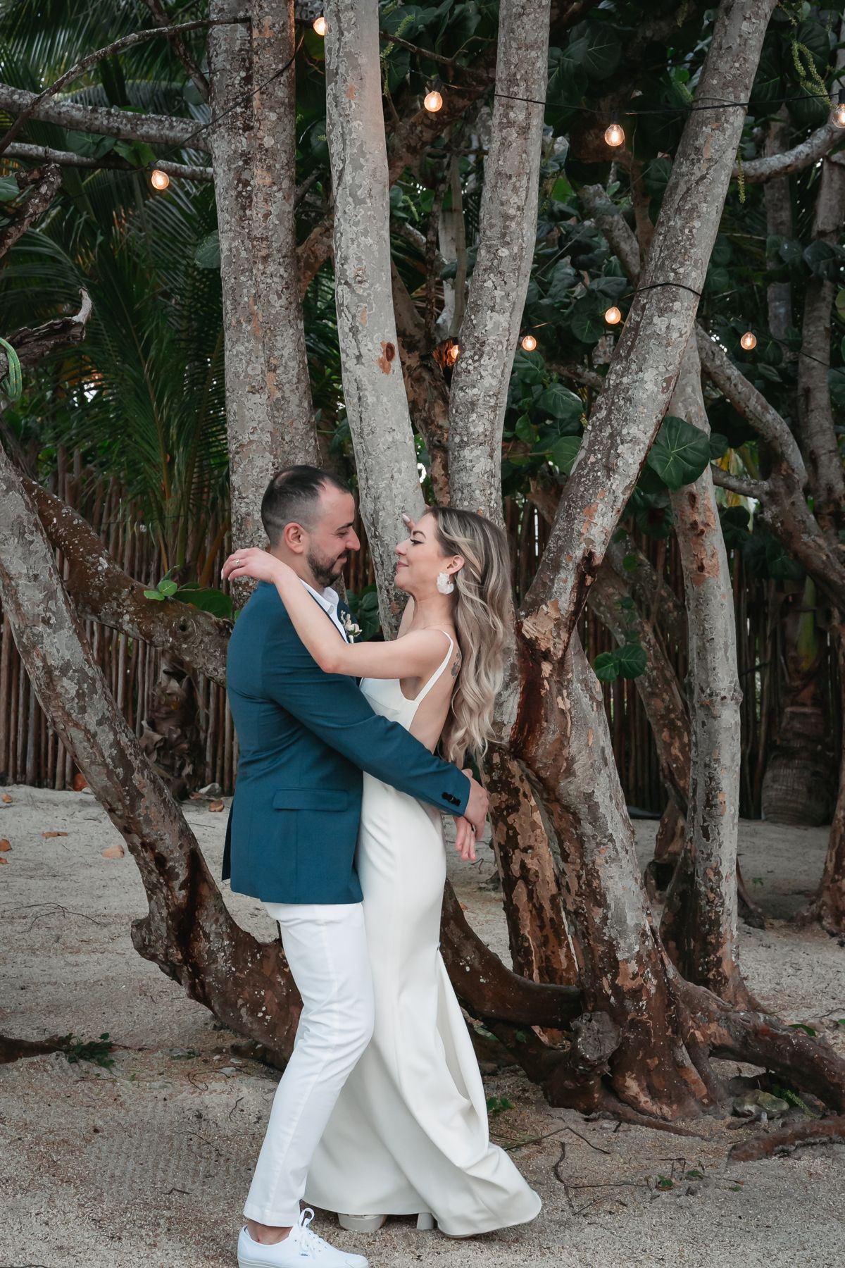 Bride and groom embrace romantically among palm trees with string lights at beach wedding.