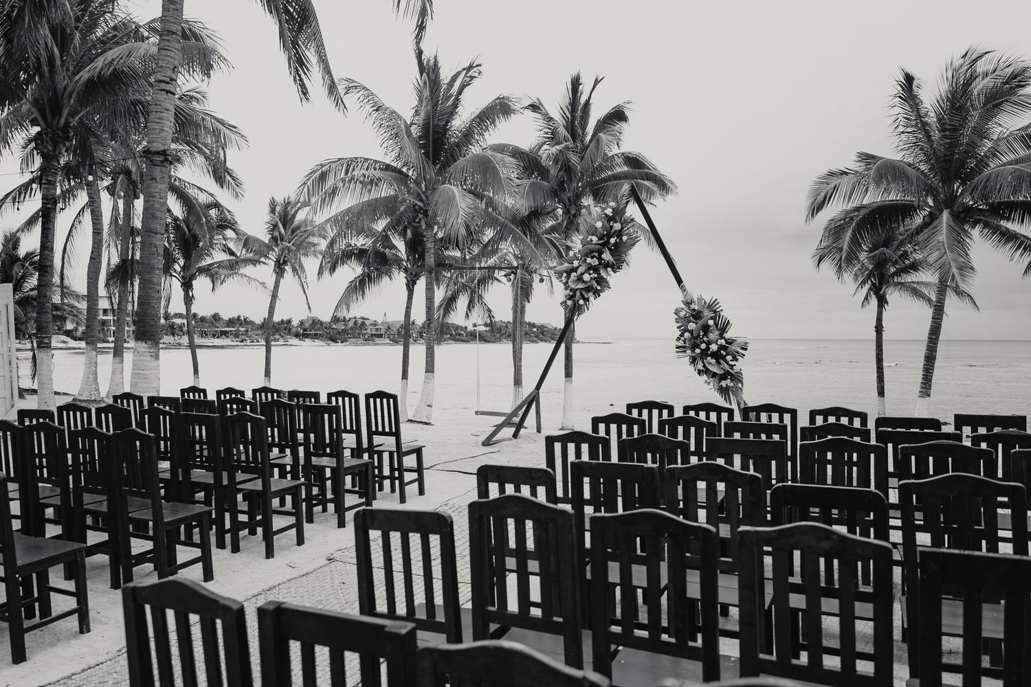 Black and white photo of a beachside wedding ceremony with rows of chairs, palm trees, and a floral arch by the sea.