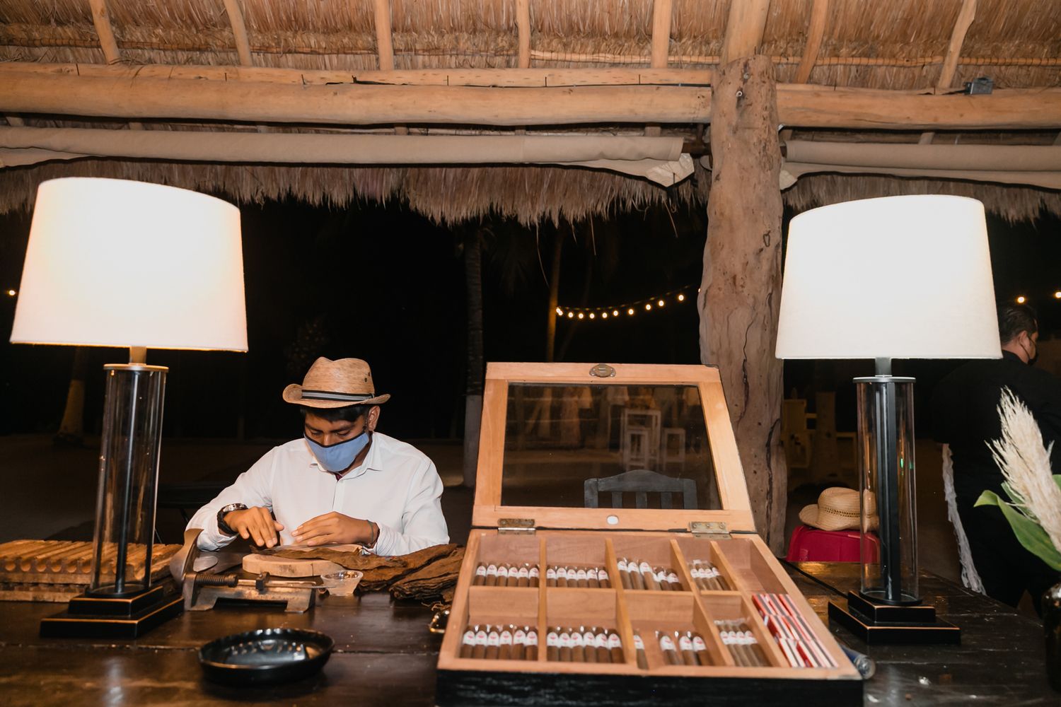 Cigar roller in hat crafts cigars at elegant display table under thatched roof at tropical wedding reception.