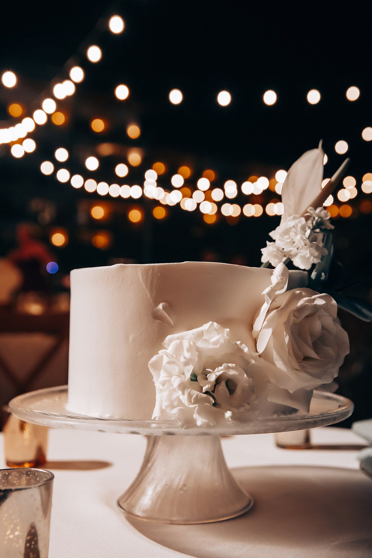 Elegant white wedding cakes displayed under romantic string lights at evening reception.