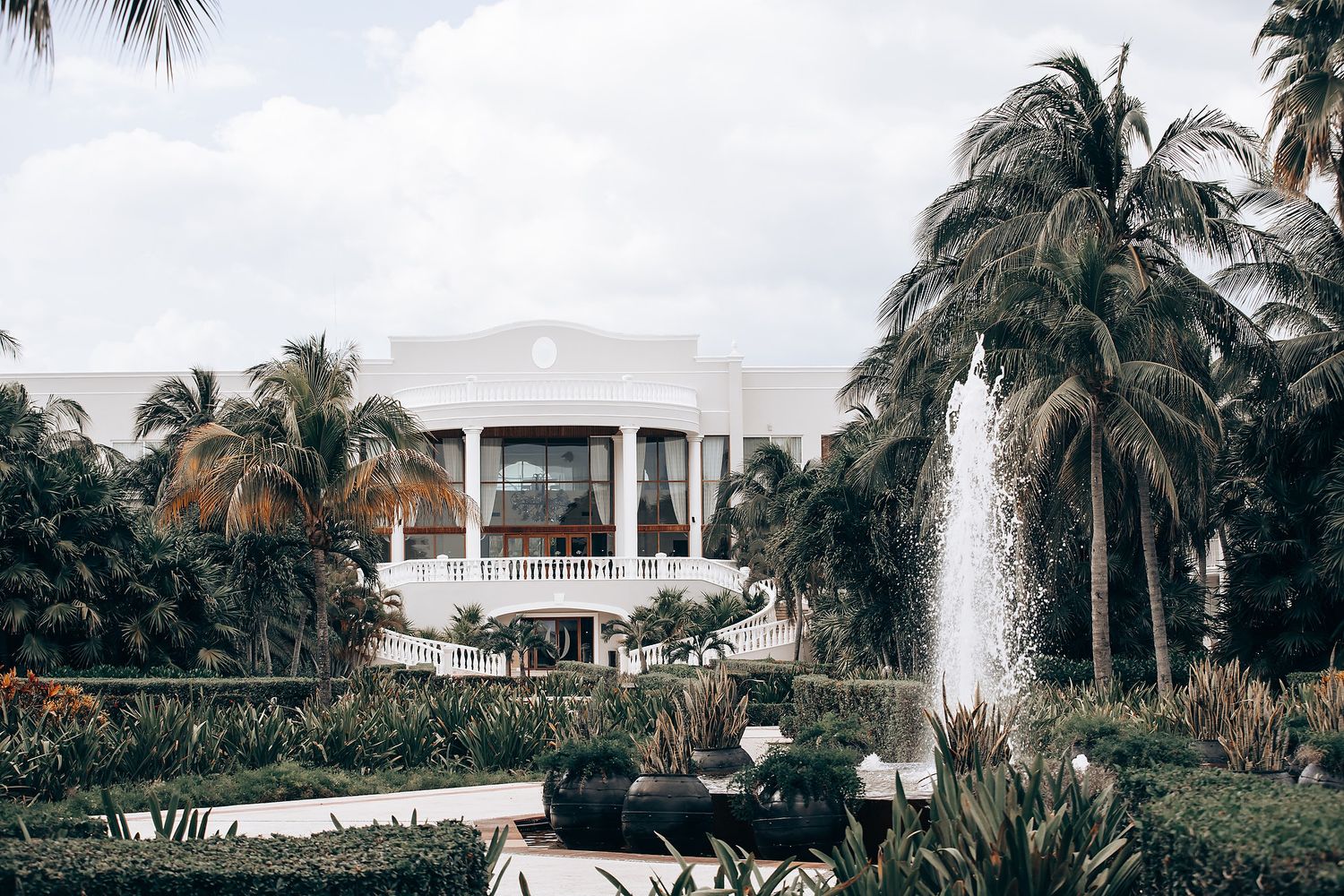 Tropical resort landscape with palm trees, fountains and white buildings along a beachfront property.