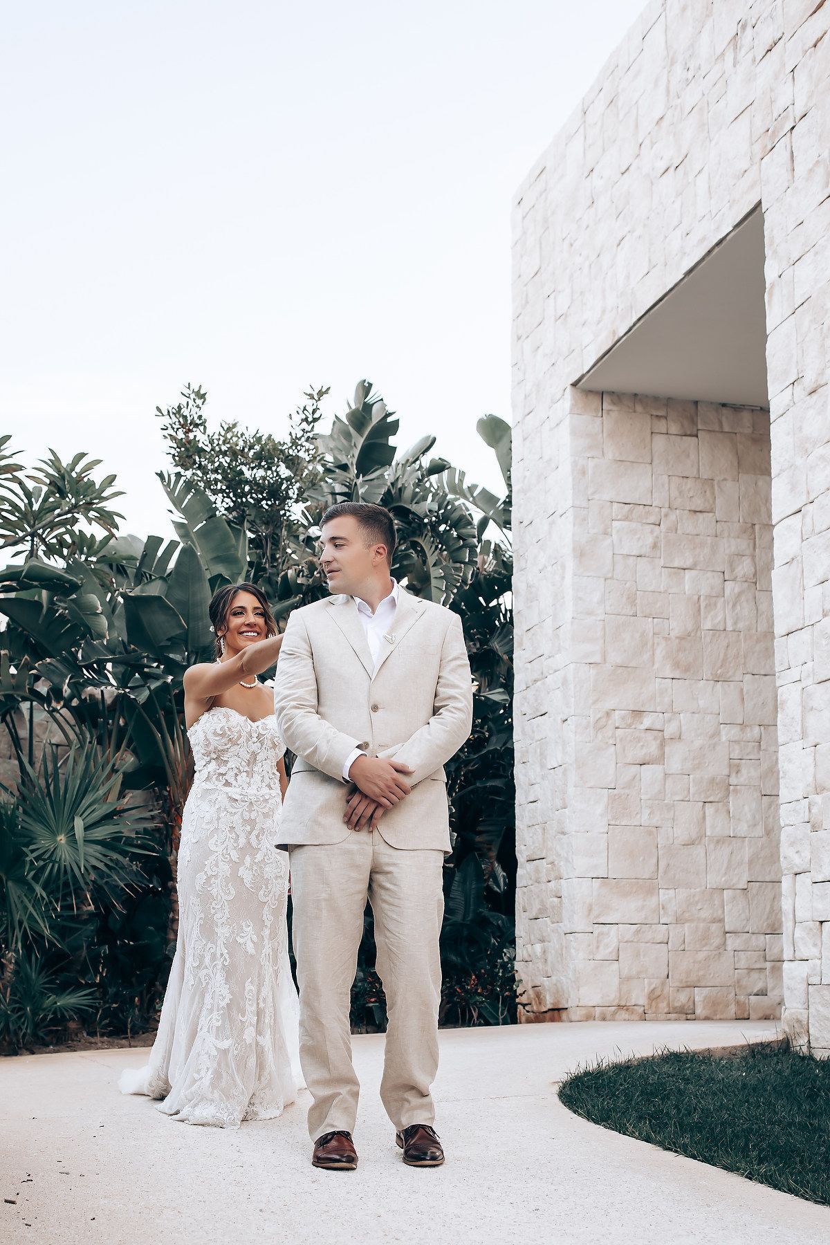 Elegant beach wedding couple poses against white stone wall surrounded by tropical greenery.