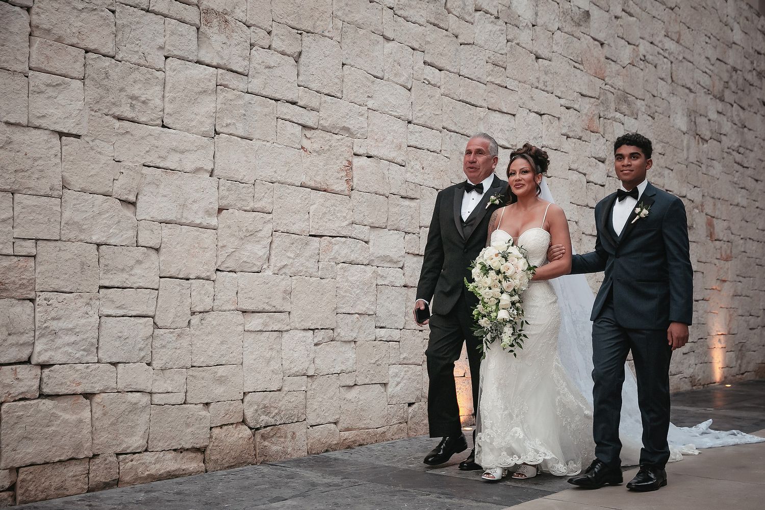 Wedding party poses against a modern stone wall backdrop during formal photography session.
